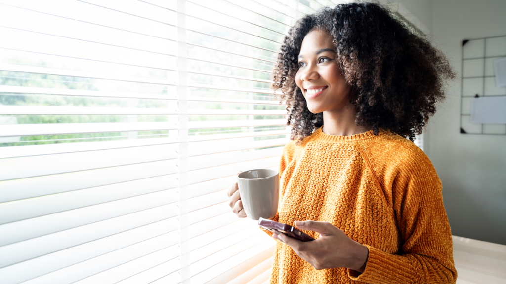 A young woman holding a coffee mug and smartphone gazes out a window, representing the self-directed digital search experience reshaping authentic multifamily marketing.